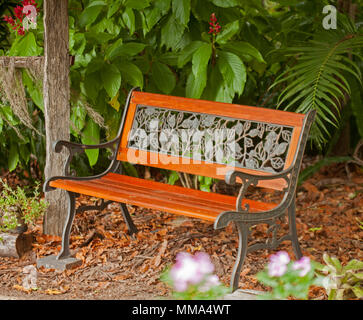 Giardino attraente banco con nero decorativo in ferro battuto e i pannelli di telaio e golden brown assicelle di legno accanto a arbusti con vivido verde fogliame Foto Stock