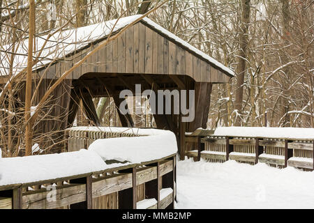 Scena invernale con avvolgimento di una passerella in legno che conduce ad un ponte coperto che attraversa un fiume in un parco boscoso. Foto Stock