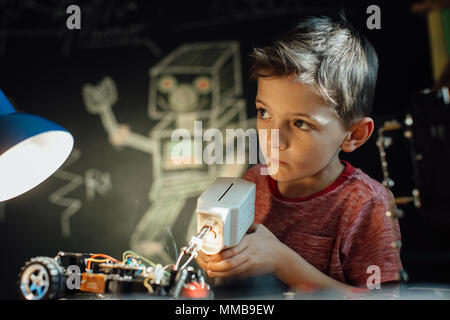 Riflessivo ragazzo lavorando sul suo progetto elettronico con una pistola per saldatura. Smart scolaro la saldatura di parti metalliche del suo giocattolo auto insieme a casa. Foto Stock