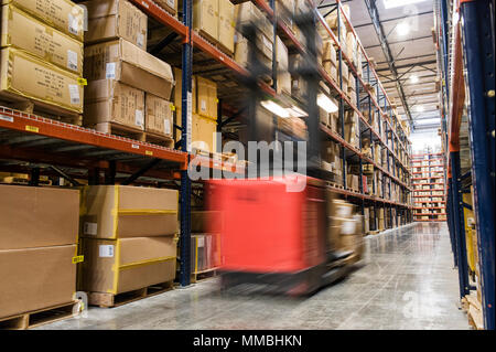 Di sfocatura di un magazzino motorizzato picker tra i corridoi di scatole di cartone su pallet impilati su scaffalature di grandi dimensioni in un grande magazzino di distribuzione. Foto Stock