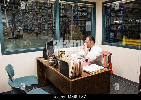 America Ispanica dirigenti che lavorano su un computer in un ufficio nel mezzo di un grande magazzino di distribuzione piena di cremagliere di scatole di cartone su pal Foto Stock