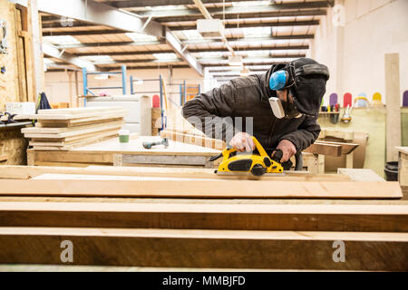 Uomo di indossare protezioni per le orecchie, occhiali protettivi e una maschera antipolvere in piedi in un magazzino, tavole di levigatura di legno riciclato. Foto Stock