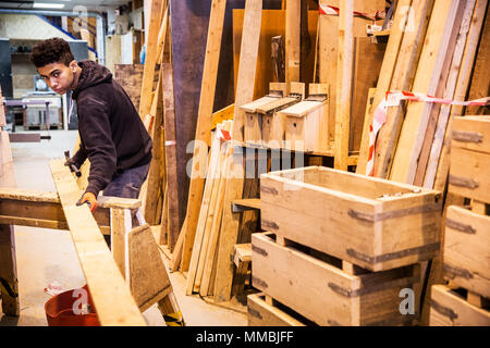 Giovane uomo che indossa guanti da lavoro in piedi in magazzino, bilanciamento listone in legno sul traliccio, la pila di carta riciclata per casse di legno. Foto Stock