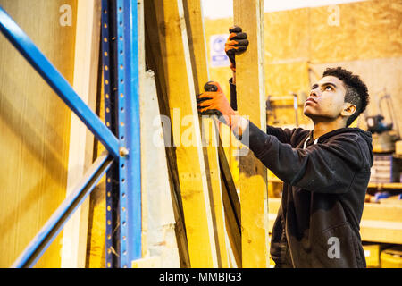 Giovane uomo che indossa guanti da lavoro in piedi accanto a una pila di assi di legno in un magazzino, tenendo plank, guardando verso l'alto. Foto Stock