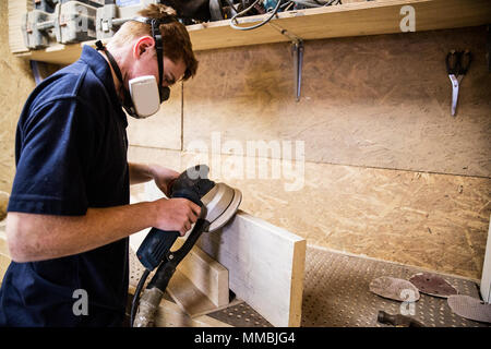 Giovane uomo che indossa la maschera antipolvere e occhiali protettivi in piedi in un workshop, utilizzando levigatrici per spianare il bordo di un pezzo di legno. Foto Stock