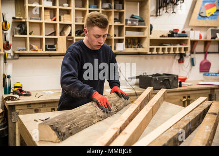 Uomo che indossa guanti da lavoro in piedi su un banco da lavoro in officina, corteccia di levigatura di un pezzo di tronco di albero. Foto Stock