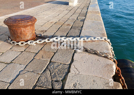 Ferro vecchio bollard con attaccato il metallo di collegamento a catena e parafango grande causando danni alla pietra istriana quay al bordo della laguna veneziana, Castell Foto Stock