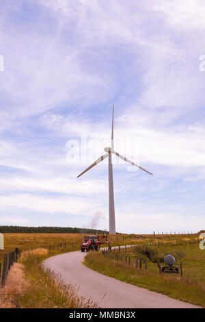 Agricoltore Homeward Bound dopo la raccolta del fieno al tramonto nel suo trattore con rastrello passando una turbina eolica fornendo renewble energia alternativa e electrici Foto Stock