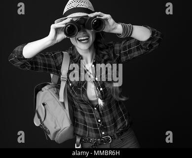 La ricerca di luoghi ispiratrice. Sorridendo turistica attiva donna con zaino guardando attraverso il binocolo isolati su Foto Stock