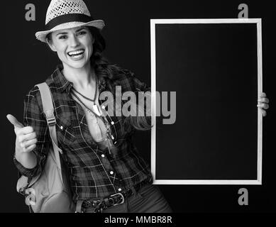 La ricerca di luoghi ispiratrice. sorridente donna attiva escursionista con zaino che mostra pollice in alto e che mostra la scheda in bianco su sfondo Foto Stock