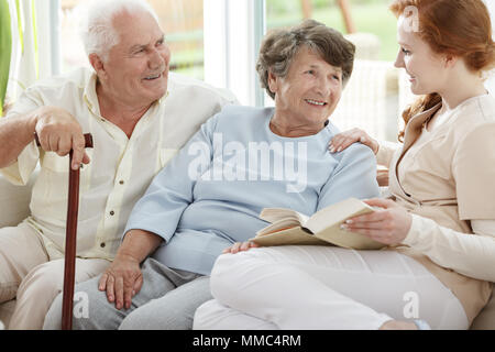 Due anziani sono guardando sorridente infermiera che legge libro in sala comune Foto Stock