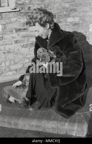 Degli anni Cinquanta, elegante signora che indossa una pelliccia inginocchiati e accarezzare il suo cane, un bassotto o salsiccia cane, tenendo nel suo braccio di altri tre nuovi cuccioli, Inghilterra, Regno Unito. Foto Stock