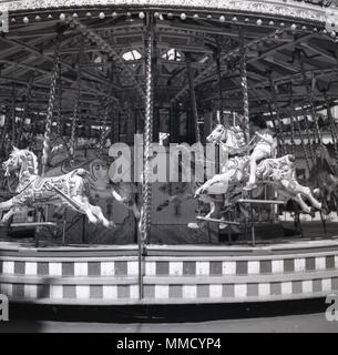 Anni sessanta, storico, giovane ragazza in sella ad un cavallo di legno su di una giostra o merry-go-round alla fiera del divertimento al parco di Battersea, Londra, Inghilterra, Regno Unito. La fiera è stata costruita per il 1951 Festival di Bretagna celebrazioni nella sezione settentrionale del parco ed è stata una delle principali attrazioni per molti anni. Foto Stock