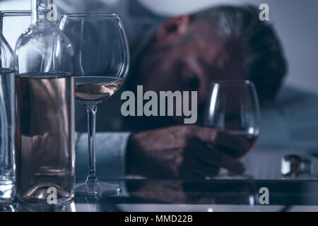 Close-up di un ubriaco disperato uomo di mezza età appoggiata sul tavolo dietro le bottiglie e i bicchieri con alcol Foto Stock