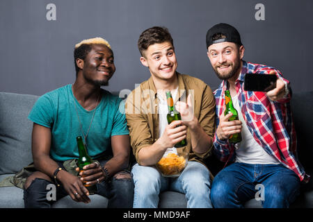 Ritratto di elegante, migliore, attraente e moderno ragazzi positivo con l'acconciatura, setole seduta sul lettino, avente tazze con lager in mani, tiro selfie Foto Stock