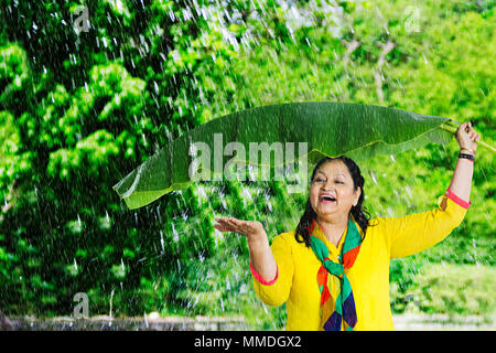 Felice Senior donna sotto Banana-Leaf mettendo mano a sentire le gocce di pioggia sul palmare Palm Foto Stock