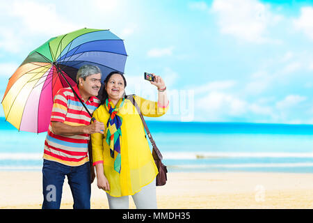 Felice coppia Senior sotto ombrellone tenendo la fotocamera Selife-Photo In-Beach vacanza vacanze Foto Stock