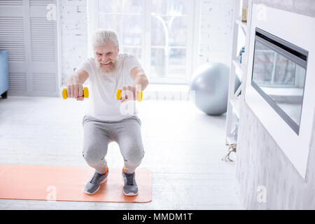 Bel uomo anziano holding manubri e facendo squat Foto Stock
