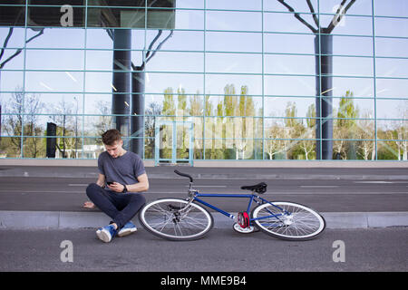 Giovani hipster utilizzare cellulare, fixed gear bike su strada di città. grande specchio background windows, urban ciclista Foto Stock