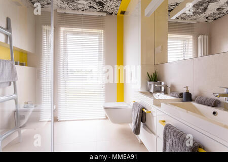 Fresco e contemporaneo luminoso bagno con mattonelle beige, specchio, graphic soffitto, doppio lavabo, wc, decorazioni in giallo e i ciechi della finestra Foto Stock