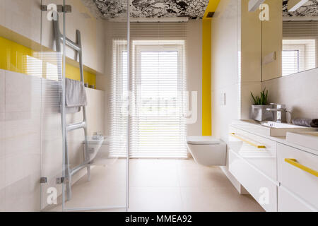Il lusso di un bagno elegante con armadi bianchi, specchio, ladder rack tower, finestra, porta in vetro e dettagli di colore giallo Foto Stock