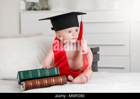 Carino baby boy in malta board hat seduta accanto alla pila di grandi libri antichi Foto Stock