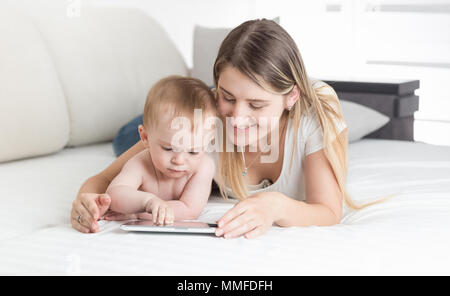 Ritratto di giovane sorridente madre Bimbo giacente sul letto e con tavoletta digitale Foto Stock