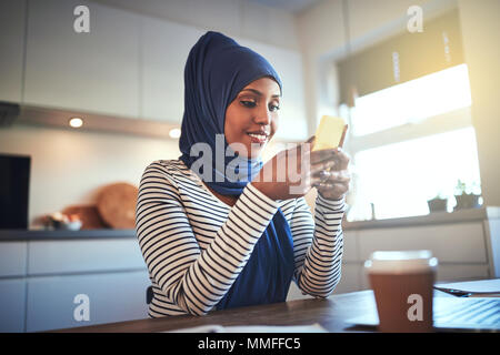 Sorridente giovani arabi donna che indossa un hijab inviando un messaggio di testo e utilizzando un computer portatile mentre è seduto nella sua cucina a casa Foto Stock