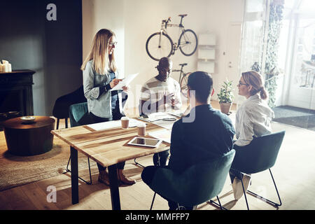 Giovane donna dando una presentazione ai colleghi seduti insieme attorno a un tavolo in un ufficio moderno Foto Stock