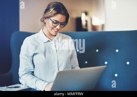 Sorridente giovane imprenditrice seduta su un divano e utilizzando un computer portatile mentre si lavora da casa Foto Stock