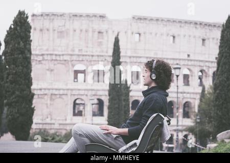Bel giovane nei panni dello sport ascoltando musica seduto su una panchina con il Colosseo in background. Giovane atleta in tuta rilassante dopo Foto Stock
