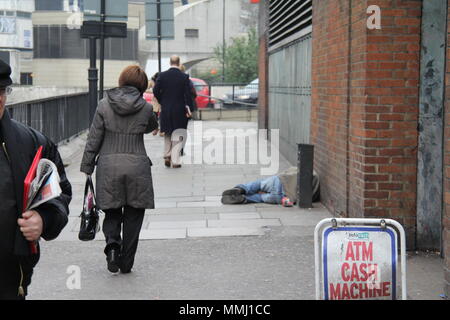 Senzatetto sonno agitato da un Bancomat firmare con la gente camminare passato. Foto Stock