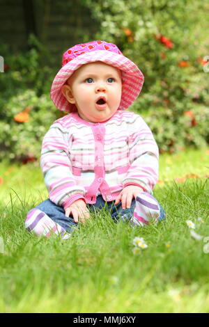 Un espressivo grande blu eyed baby siede sul prato con un cappello e abito luminoso, circondato da fiori. Foto Stock