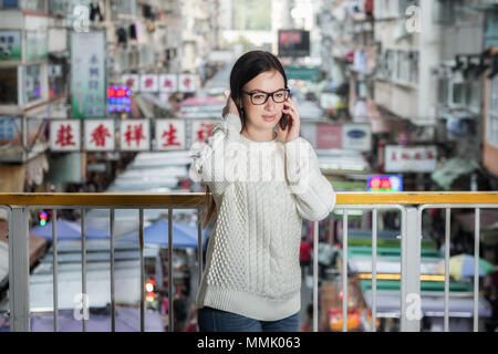 Attraente sorridente brunette donna in occhiali permanente sulla passerella pedonale e parlare per telefono cellulare. Hong Kong street market sullo sfondo. Foto Stock