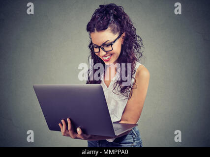 Casual giovane donna shopping online utilizzando laptop e sorridendo allegramente sul muro grigio Sfondo. Foto Stock