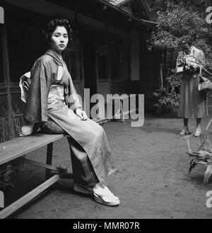 Degli anni Cinquanta, storico attraente giovane femmina adulta seduta su una panchina al di fuori di indossare l'abbigliamento tradizionale giapponese o di costume, il kimono e fotografata da un western lady. Il kimono è un vestito di seta che viene avvolto attorno al corpo, legata con un nastro largo, un obi e è il costume nazionale per il Giappone. Foto Stock