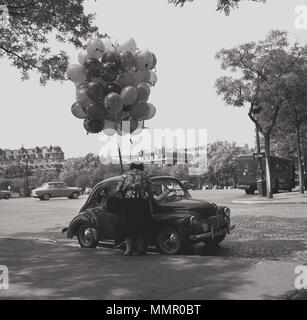 1950s, storica, una piccola auto dell'epoca parcheggiata su una strada acciottolata accanto ad un marciapiede che acquista balloni da una donna di strada parigina commerciante, Parigi, Francia. Foto Stock