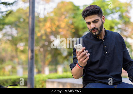 Bel giovane uomo indiano utilizzando il telefono cellulare mentre è seduto sul co Foto Stock