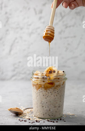 Dieta sana prima colazione. Durante la notte i fiocchi d'avena con semi di Chia, banane, burro di arachidi, miele, cioccolato spolverata in un vasetto di vetro su di un cemento grigio backg Foto Stock