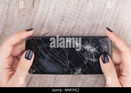 Uno smartphone con uno schermo rotto nella mano della ragazza su di un tavolo di legno. Close-up. Studio di illuminazione. Foto Stock