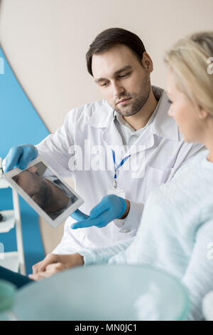 Uomo Barbuto in studio dentistico che mostra il tablet PC alla giovane cliente avente la consultazione in armadio. Foto Stock