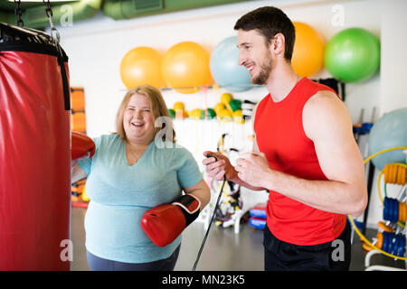 Ritratto di felice donna obesa colpire sacco da boxe e ridere mentre ti alleni in palestra con personal fitness coach Foto Stock