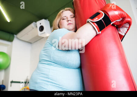 Angolo basso ritratto di esaurito donna obesa appoggiata sul sacco da boxe con gli occhi chiusi dopo il calo ponderale estremo della formazione in palestra, spazio di copia Foto Stock