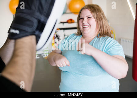 Vita ritratto di sorridere donna obesa che lavora fuori in palestra con personal fitness coach, colpire i pad Foto Stock
