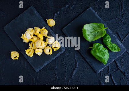 Preparato di fresco in casa materie flouring tortellini pronti a cuocere sulla pietra scura la piastra con le foglie di basilico su sfondo nero. La cucina tradizionale italiana Foto Stock