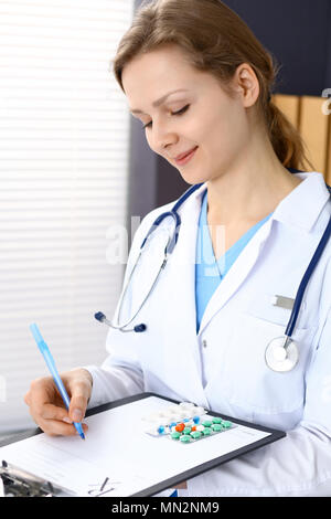 Donna medico prescrizione di scrittura mentre è seduto in ufficio dell'ospedale. Un sacco di varie pillole colorate liye presso i suoi appunti. Medicina e salute Foto Stock