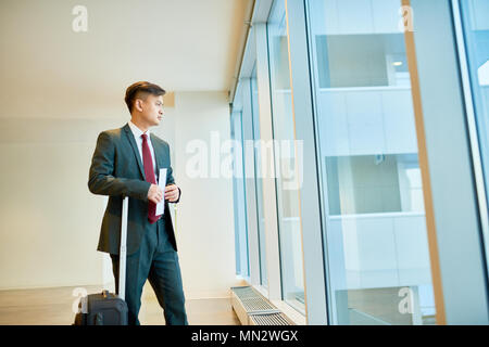 Vista laterale ritratto di successo imprenditore asiatica guardando alla finestra in aeroporto in attesa per il volo con i biglietti e la valigia, spazio di copia Foto Stock