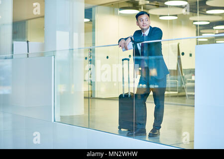 A piena lunghezza ritratto pf moderno uomo asiatico indossando business suit in piedi alla finestra in aeroporto in attesa del volo con il bagaglio e spazio di copia Foto Stock