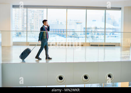 Vista laterale verticale del moderno imprenditore asiatici valigetta e parlare al telefono mentre si incrociano airport lounge di attesa, spazio di copia Foto Stock