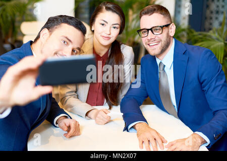 Ritratto di tre giovani uomini di affari tenendo gruppo selfie tramite smartphone mentre è seduto al tavolo del bar durante la pausa caffè Foto Stock